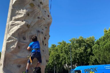Enfant grimpant sur un mur d'escalade dans un parc, sous le regard d'un animateur qui l'assure, lors du Prox Raid Aventure qui s'est déroulé au parc du Grenouillet à Cavaillon en juillet 2022