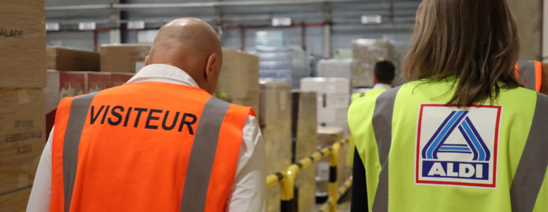 Deux personnes de dos vêtue d'un gilet jaune et orange réfléchissant marchant dans un entrepôt logistique de Cavaillon