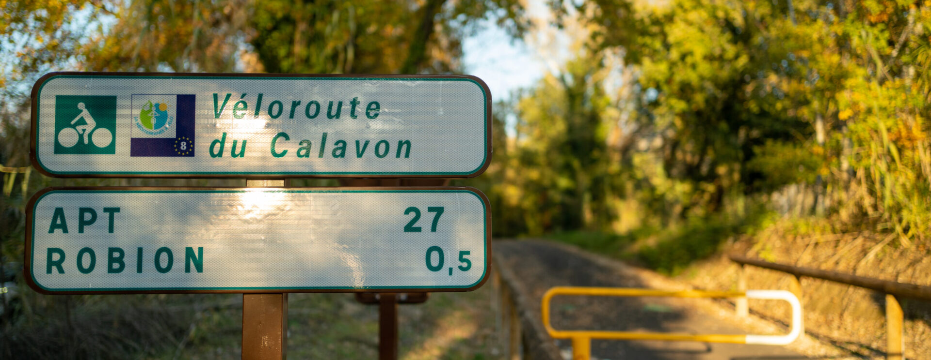 Photo d'illustration de la Véloroute du Calavon montrant un panneau de signalisation indiquant qu'Apt est à 27km et Robion à 500 mètres avec pour fond la véloroute.
