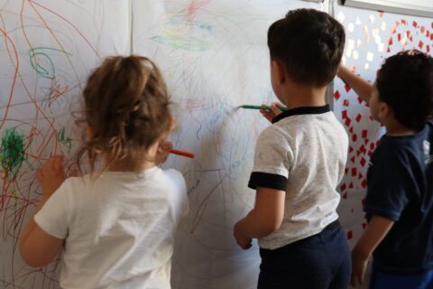 Trois jeunes enfants de dos (une fille et deux garçons) en train de dessiner sur une feuille accrochée sur un mur, en crèche.