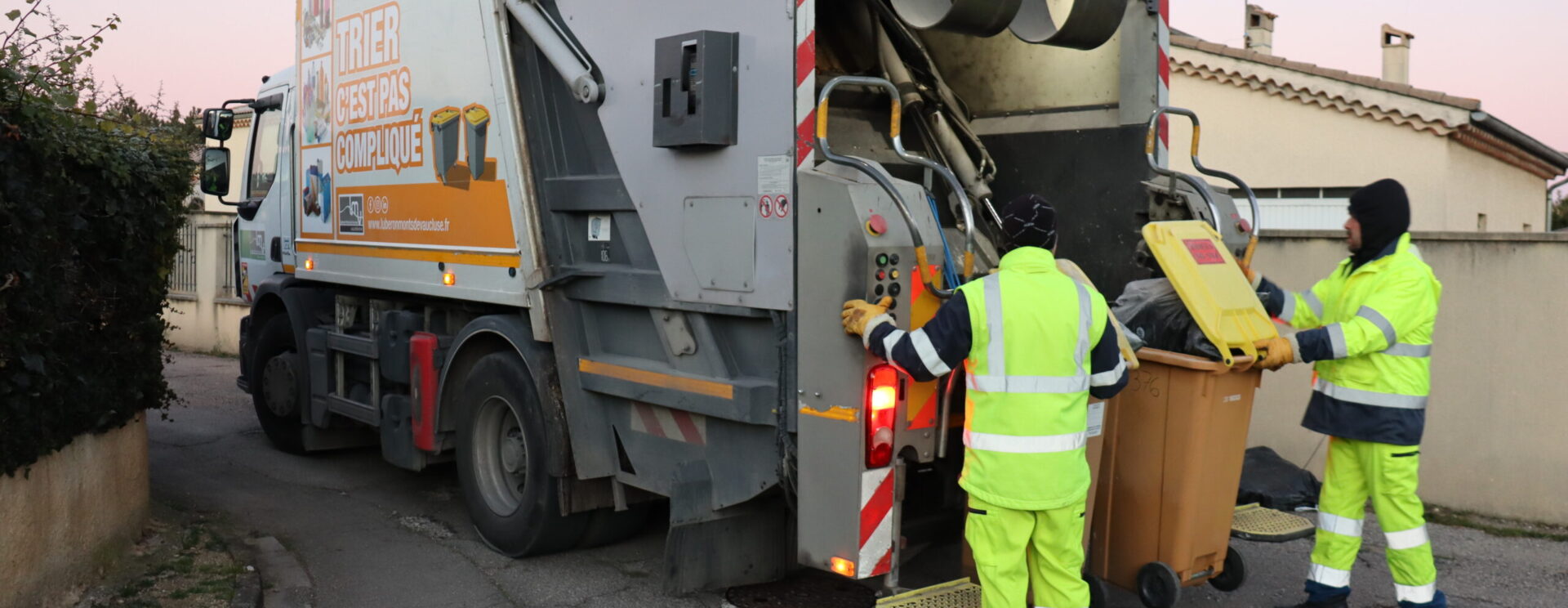 Deux agents de collecte LMV habillés en tenue de sécurité jaune fluo en train de vider un bac roulant de déchets dans un camion de collecte