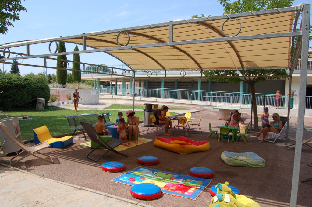Coin lecture du centre aquatique de plein air de Cavaillon, équipé de transats, tapis de sols, coussins, petites tables et chaises pour enfant. Le lieu est ombragé par une tonnelle. Des personnes (adultes et enfants), profitent du lieu en lisant ou dessinant. - Agrandir l'image, fenêtre modale