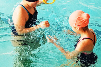 Deux enfants, bonnet de piscine sur les yeux, dans le petit bassin de la piscine, en plein cours de sensibilisation au handicap (ici visuel) - Agrandir l'image 5 sur 6, fenêtre modale