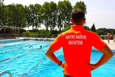 Maître-nageur debout les bras sur les hanches en train de surveiller un bassin dans lequel des personnes sont en train de nager